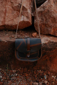 A leather bag with a flap closure is hanging against a backdrop of large, rugged rocks. The bag has a simple design and a natural brown tone, blending with the earthy surroundings of rocky, uneven ground.