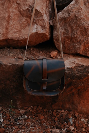 A leather bag with a flap closure is hanging against a backdrop of large, rugged rocks. The bag has a simple design and a natural brown tone, blending with the earthy surroundings of rocky, uneven ground.