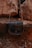A leather bag with a flap closure is hanging against a backdrop of large, rugged rocks. The bag has a simple design and a natural brown tone, blending with the earthy surroundings of rocky, uneven ground.