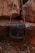 A leather bag with a flap closure is hanging against a backdrop of large, rugged rocks. The bag has a simple design and a natural brown tone, blending with the earthy surroundings of rocky, uneven ground.