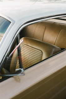 A close-up view of the interior of a car featuring tan leather seats with horizontal stitching. The image captures part of the window and the side mirror, reflecting natural light.
