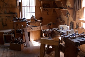 A traditional woodworking shop with a variety of tools and benches, lit by natural light streaming through windows. Tools such as saws, hammers, and chisels are organized on shelves and wooden benches, and a wooden floor is covered with wood shavings.
