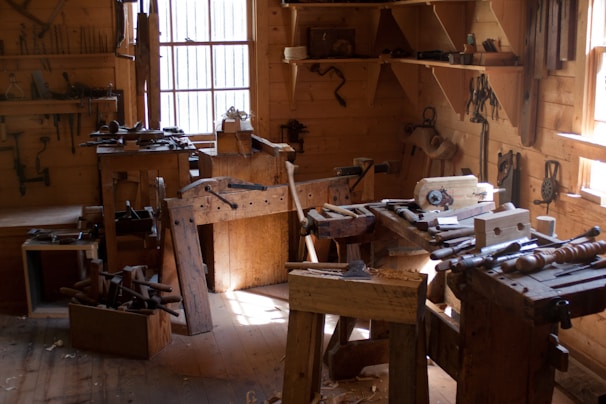A traditional woodworking shop with a variety of tools and benches, lit by natural light streaming through windows. Tools such as saws, hammers, and chisels are organized on shelves and wooden benches, and a wooden floor is covered with wood shavings.