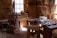 A traditional woodworking shop with a variety of tools and benches, lit by natural light streaming through windows. Tools such as saws, hammers, and chisels are organized on shelves and wooden benches, and a wooden floor is covered with wood shavings.