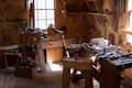 A traditional woodworking shop with a variety of tools and benches, lit by natural light streaming through windows. Tools such as saws, hammers, and chisels are organized on shelves and wooden benches, and a wooden floor is covered with wood shavings.
