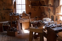A traditional woodworking shop with a variety of tools and benches, lit by natural light streaming through windows. Tools such as saws, hammers, and chisels are organized on shelves and wooden benches, and a wooden floor is covered with wood shavings.
