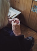 An elderly individual is seen sitting in a cozy, wood-paneled room. They are holding a small container of pinkish cream, gently applying it with their fingers. The focus is on their hands, showing the texture of their skin, suggesting age.