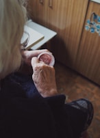 An elderly individual is seen sitting in a cozy, wood-paneled room. They are holding a small container of pinkish cream, gently applying it with their fingers. The focus is on their hands, showing the texture of their skin, suggesting age.