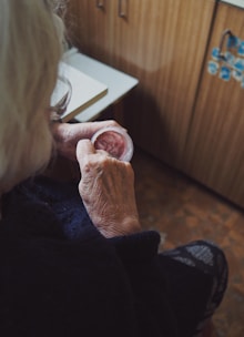 An elderly individual is seen sitting in a cozy, wood-paneled room. They are holding a small container of pinkish cream, gently applying it with their fingers. The focus is on their hands, showing the texture of their skin, suggesting age.