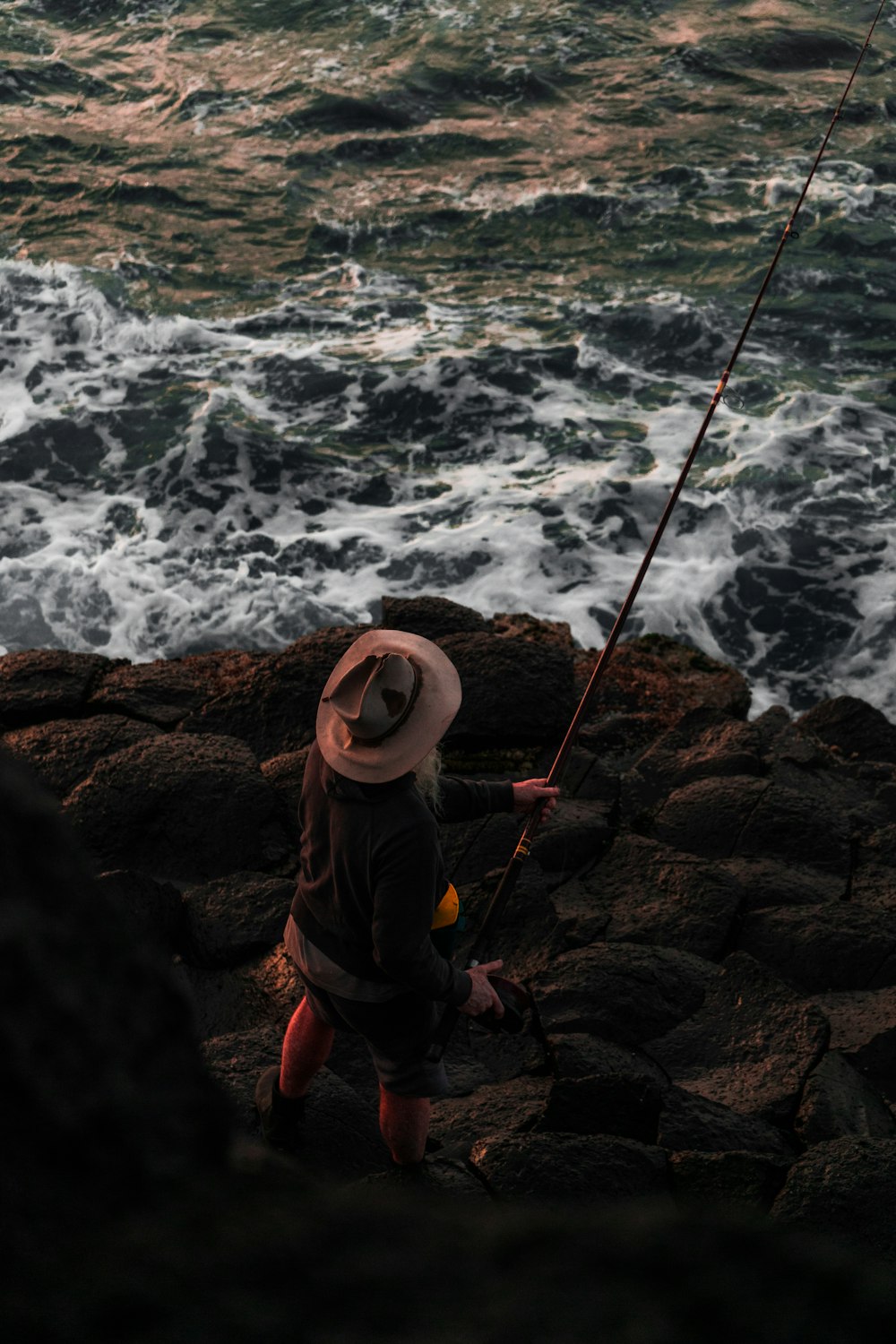 man in brown hat and yellow jacket sitting on rock near sea waves crashing on shore