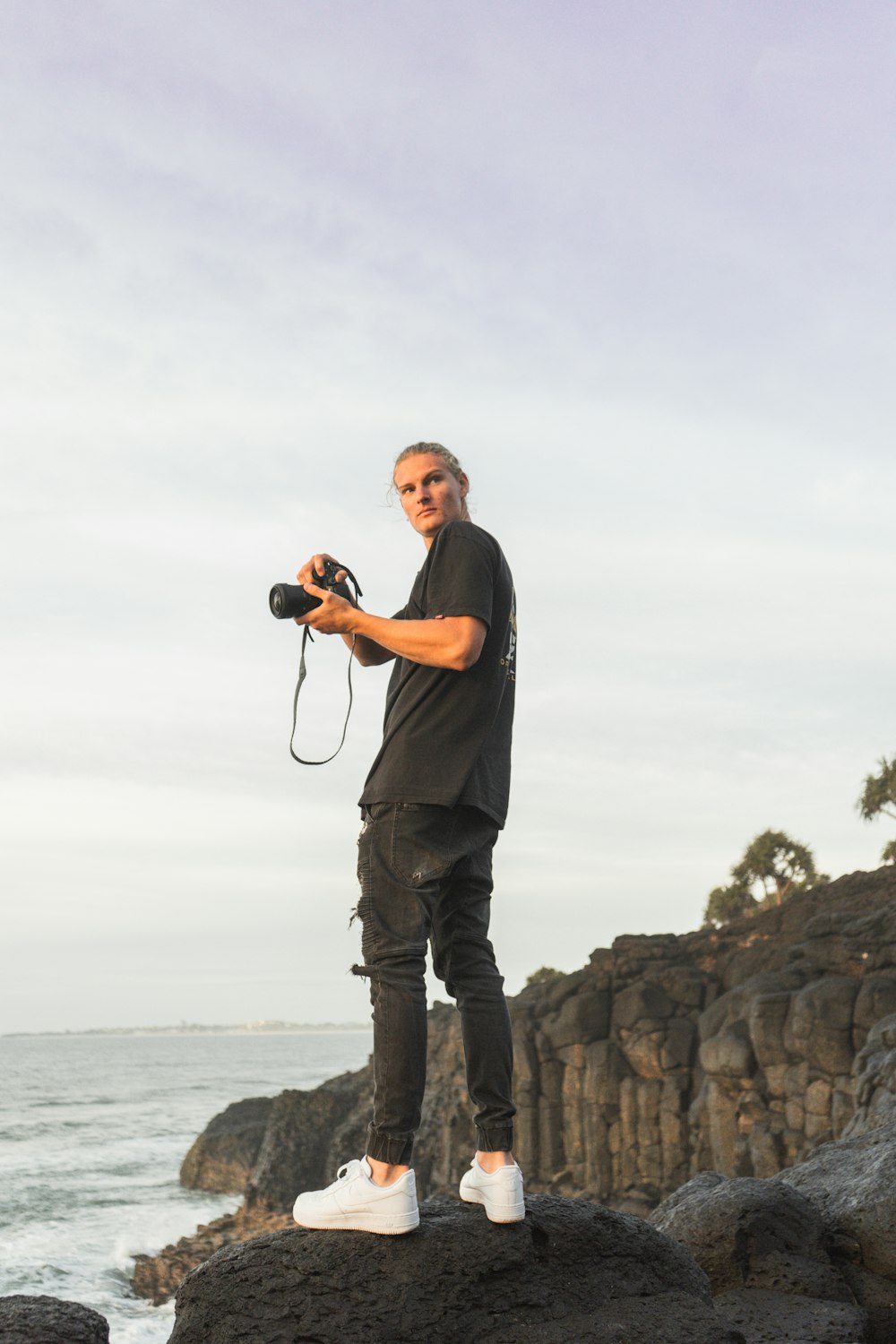 man in black t-shirt and gray pants holding camera standing on rock formation near sea