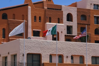 A vibrant hotel facade with earthy tones of brown and orange is captured, featuring architectural details such as rounded windows and multiple balconies. In front of the building, three flags are prominently displayed: a white flag with a logo, the Mexican flag, and the United States flag. The sky is clear and blue, adding to the brightness of the scene.