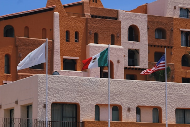 A vibrant hotel facade with earthy tones of brown and orange is captured, featuring architectural details such as rounded windows and multiple balconies. In front of the building, three flags are prominently displayed: a white flag with a logo, the Mexican flag, and the United States flag. The sky is clear and blue, adding to the brightness of the scene.