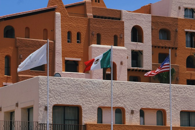A vibrant hotel facade with earthy tones of brown and orange is captured, featuring architectural details such as rounded windows and multiple balconies. In front of the building, three flags are prominently displayed: a white flag with a logo, the Mexican flag, and the United States flag. The sky is clear and blue, adding to the brightness of the scene.
