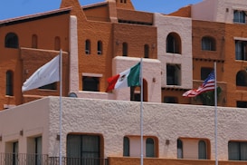 A vibrant hotel facade with earthy tones of brown and orange is captured, featuring architectural details such as rounded windows and multiple balconies. In front of the building, three flags are prominently displayed: a white flag with a logo, the Mexican flag, and the United States flag. The sky is clear and blue, adding to the brightness of the scene.
