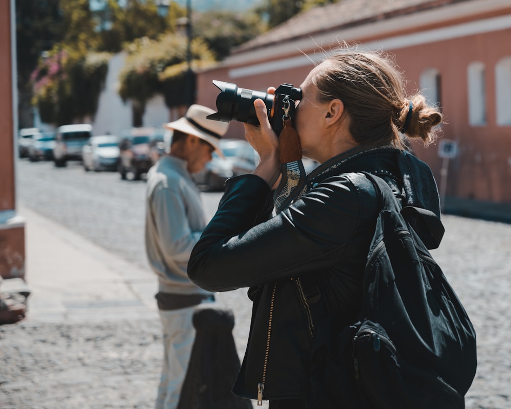 Mujer con chaqueta de cuero negro usando cámara DSLR negra durante el día