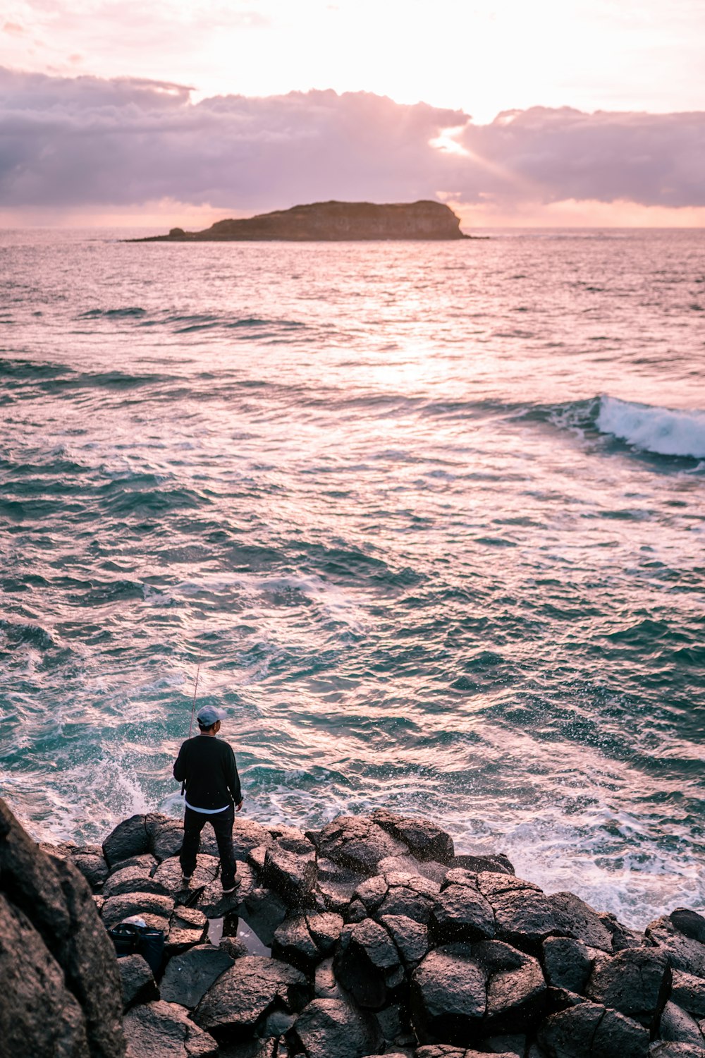 Mann in schwarzer Jacke steht tagsüber auf einer Felsformation in der Nähe eines Gewässers