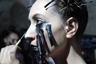 A person is having dark, dramatic makeup applied to their face. Black lines resembling tears extend down from heavily shadowed eyelids, while their hair is styled with numerous bobby pins holding it in place.