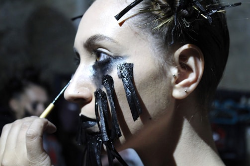 A person is having dark, dramatic makeup applied to their face. Black lines resembling tears extend down from heavily shadowed eyelids, while their hair is styled with numerous bobby pins holding it in place.