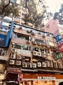 A building facade with numerous air conditioning units mounted on the exterior. The lower floors feature shop signs, including a 'mannings' store. Tall trees with green foliage are seen above, casting shadows on the building. The urban environment is characterized by high-rise structures and a mix of commercial signage.