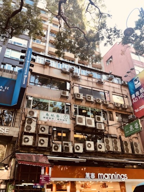 A building facade with numerous air conditioning units mounted on the exterior. The lower floors feature shop signs, including a 'mannings' store. Tall trees with green foliage are seen above, casting shadows on the building. The urban environment is characterized by high-rise structures and a mix of commercial signage.
