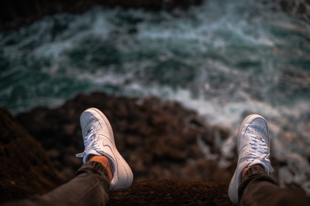 person in brown pants and white sneakers sitting on brown rock near body of water during