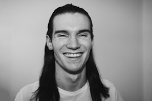 A monochrome portrait of a person with long hair, smiling broadly. The image captures the person's expression in a candid and joyful moment against a plain background.