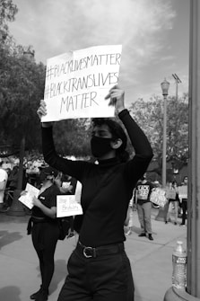 A person stands with arms raised, holding a sign with the messages '#BLACKLIVESMATTER' and '#BLACKTRANSLIVESMATTER'. Other individuals in the background hold similar protest signs, indicating participation in a social justice demonstration.
