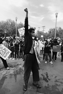 A black and white photograph captures a protest scene with a crowd of people. The central figure stands prominently at the front with one arm raised in a fist, symbolizing solidarity and resistance. The protester wears a mask, and signs with messages such as 'BLACK LIVES MATTER' are visible. The scene is set outdoors with trees and street lamps visible in the background.