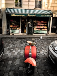 A vintage red Vespa scooter is parked on a cobblestone street in front of a small grocery store. The store has a green and white striped awning with 'Alimentation Générale' written on it, along with a phone number. The store displays various fruits and vegetables in colorful arrangements inside and outside the door.