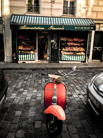 A vintage red Vespa scooter is parked on a cobblestone street in front of a small grocery store. The store has a green and white striped awning with 'Alimentation Générale' written on it, along with a phone number. The store displays various fruits and vegetables in colorful arrangements inside and outside the door.