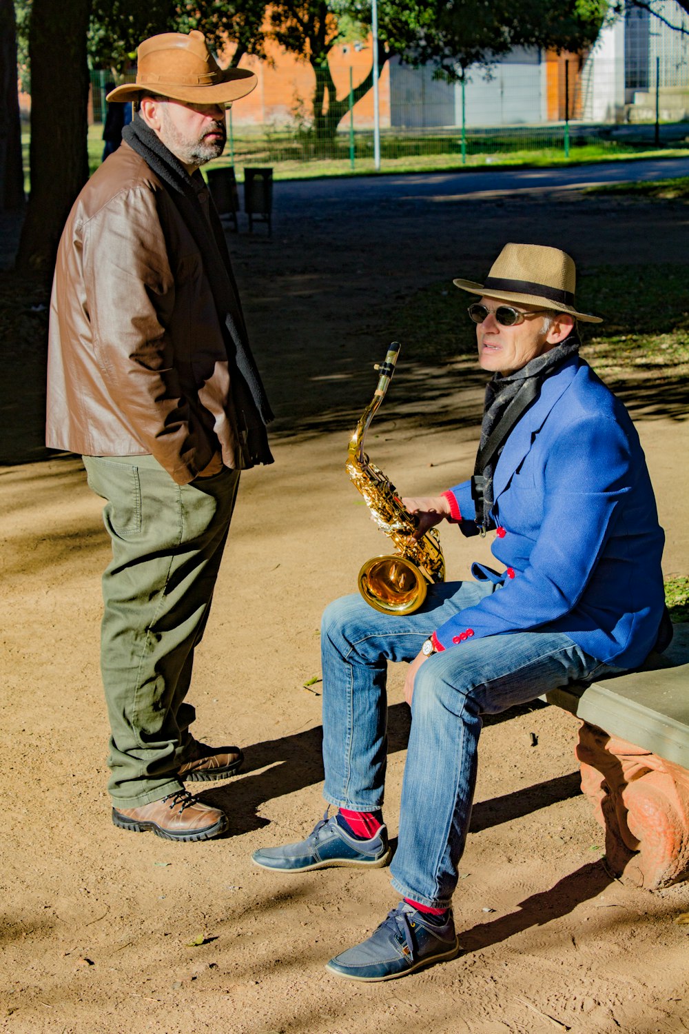man in blue jacket and blue denim jeans sitting on brown wooden bench playing saxophone