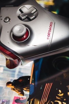 A close-up of a silver and red gaming controller with visible buttons and a joystick. The controller has a black D-pad and an 'Arasaka' logo. In the background, there is a blurred image with vibrant colors, depicting a person in a jacket and a futuristic car, suggesting a scene from a video game.