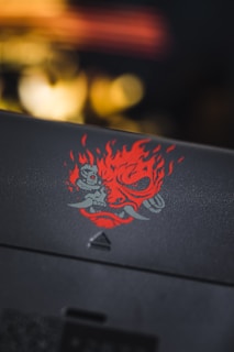 A detailed depiction of a fiery, stylized design featuring a red face with flames and tusks. The art is striking and intricate, set against a dark, blurred background.
