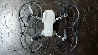 The image depicts a quadcopter drone lying on a dark, textured surface. The drone has four propellers and is equipped with propeller guards. Its body is mostly white with some black or dark gray components.