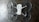 The image depicts a quadcopter drone lying on a dark, textured surface. The drone has four propellers and is equipped with propeller guards. Its body is mostly white with some black or dark gray components.