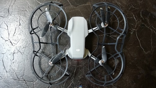 The image depicts a quadcopter drone lying on a dark, textured surface. The drone has four propellers and is equipped with propeller guards. Its body is mostly white with some black or dark gray components.