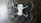 The image depicts a quadcopter drone lying on a dark, textured surface. The drone has four propellers and is equipped with propeller guards. Its body is mostly white with some black or dark gray components.