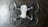 The image depicts a quadcopter drone lying on a dark, textured surface. The drone has four propellers and is equipped with propeller guards. Its body is mostly white with some black or dark gray components.