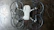 The image depicts a quadcopter drone lying on a dark, textured surface. The drone has four propellers and is equipped with propeller guards. Its body is mostly white with some black or dark gray components.