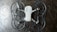 The image depicts a quadcopter drone lying on a dark, textured surface. The drone has four propellers and is equipped with propeller guards. Its body is mostly white with some black or dark gray components.