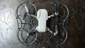 The image depicts a quadcopter drone lying on a dark, textured surface. The drone has four propellers and is equipped with propeller guards. Its body is mostly white with some black or dark gray components.