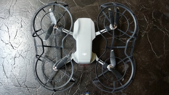 The image depicts a quadcopter drone lying on a dark, textured surface. The drone has four propellers and is equipped with propeller guards. Its body is mostly white with some black or dark gray components.