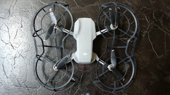 The image depicts a quadcopter drone lying on a dark, textured surface. The drone has four propellers and is equipped with propeller guards. Its body is mostly white with some black or dark gray components.