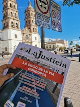 A person's hand holds a newspaper titled 'La Justicia' in front of a historic building with two towers. The newspaper features an article criticizing local government spending on infrastructure. Road signs prohibiting motorbikes, bicycles, and right turns are visible in the foreground, against a bright blue sky.