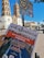 A person's hand holds a newspaper titled 'La Justicia' in front of a historic building with two towers. The newspaper features an article criticizing local government spending on infrastructure. Road signs prohibiting motorbikes, bicycles, and right turns are visible in the foreground, against a bright blue sky.