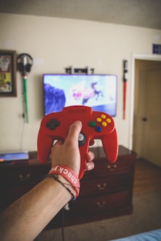 A person is holding a red retro-style video game controller in front of a television with an action game on the screen. The room has a wooden dresser below the TV and headphones hanging on the wall.