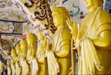 A row of yellow Buddha statues adorned with intricate patterns and decorative elements, each holding prayer beads. The background features ornate mosaic designs, adding to the spiritual atmosphere.