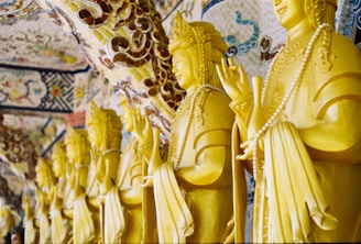 A row of yellow Buddha statues adorned with intricate patterns and decorative elements, each holding prayer beads. The background features ornate mosaic designs, adding to the spiritual atmosphere.