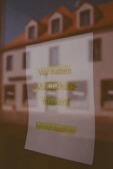 A paper sign is taped to a window displaying German text. The text reads 'Wir haben Mundschutzmasken! Bahnhof Apotheke', which translates to 'We have face masks! Station Pharmacy'. The blurred reflection of a building with a red roof is visible in the glass.